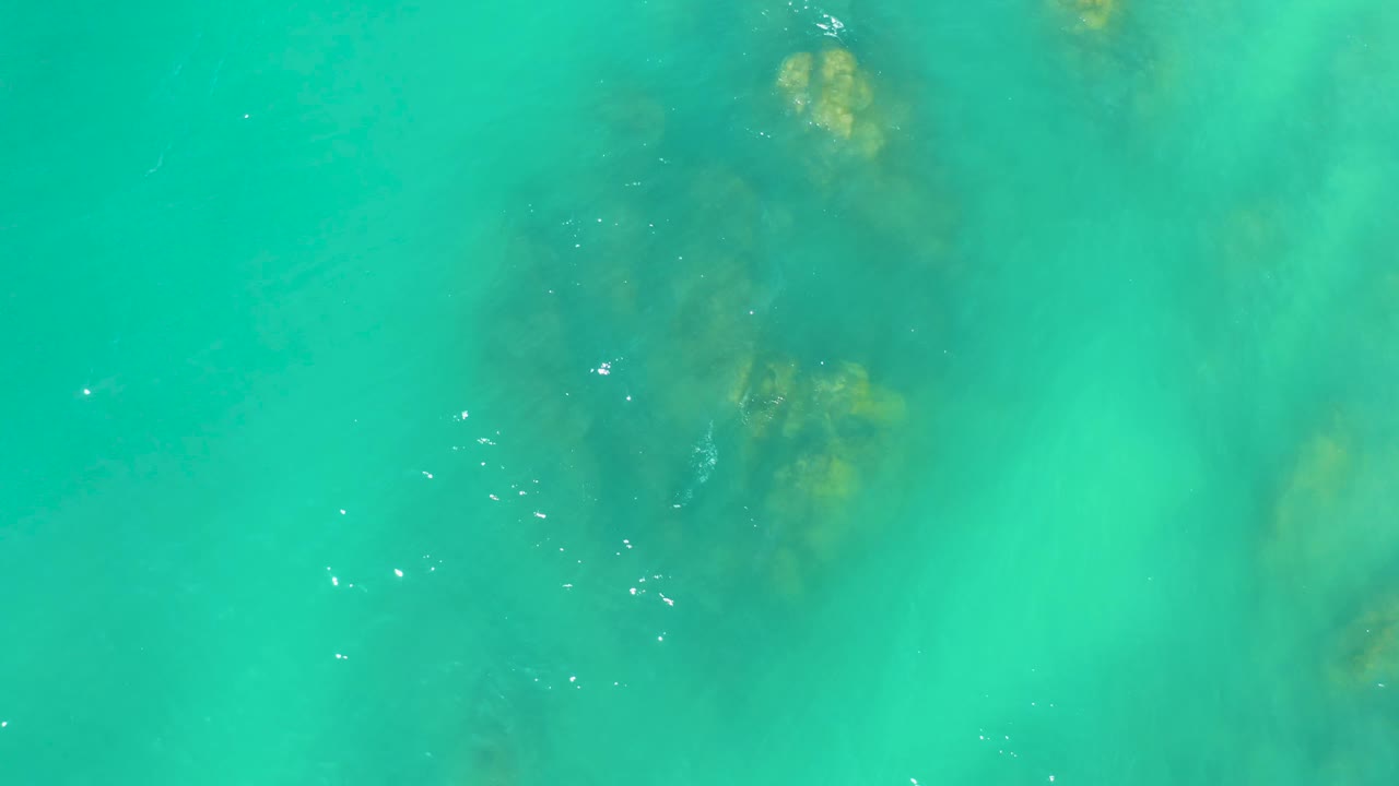 Overhead frame from Dreams Los Cabos showing transparent green-blue water, soft ripples, and a darker rock patch below the surface with no visible people.
