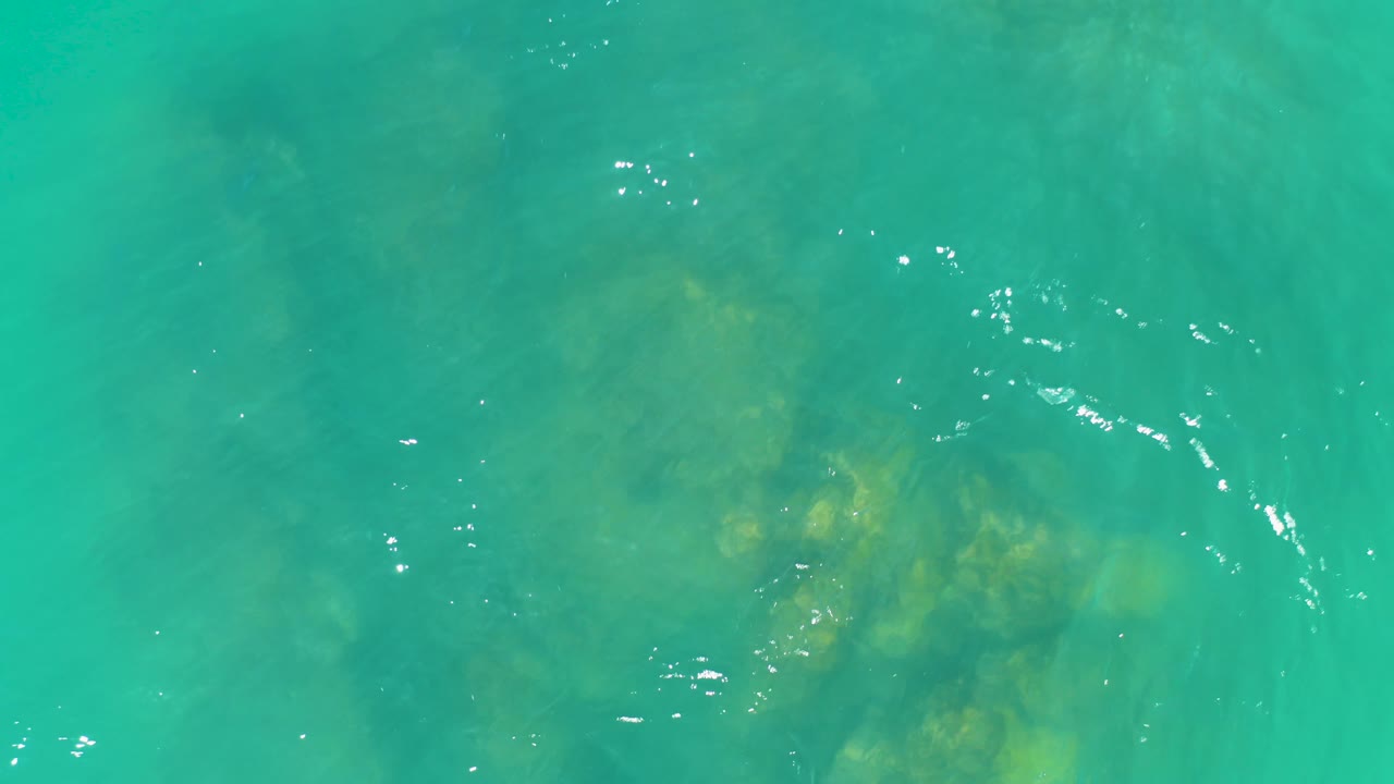 A top-down Dreams Los Cabos frame shows clustered submerged rocks or coral under clear turquoise water, with small white reflections and no visible people.