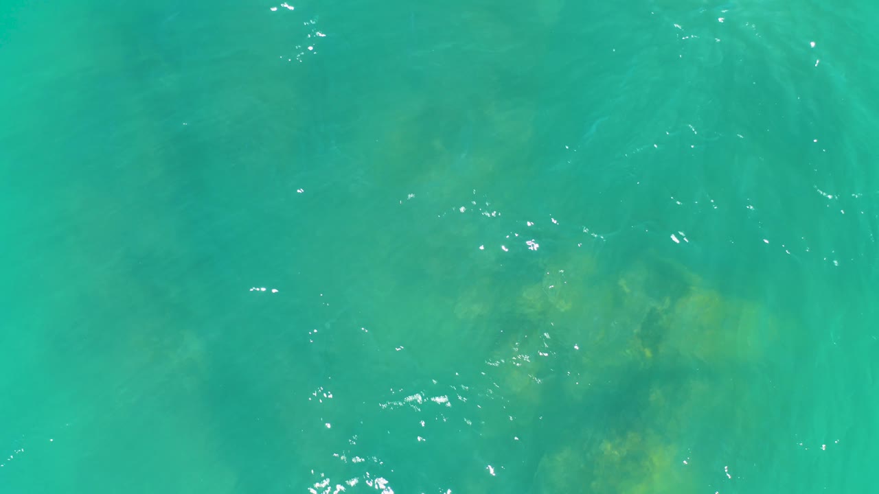 Clear turquoise water at Dreams Los Cabos reveals a darker submerged rock patch beneath soft ripples, with no visible people or structures.