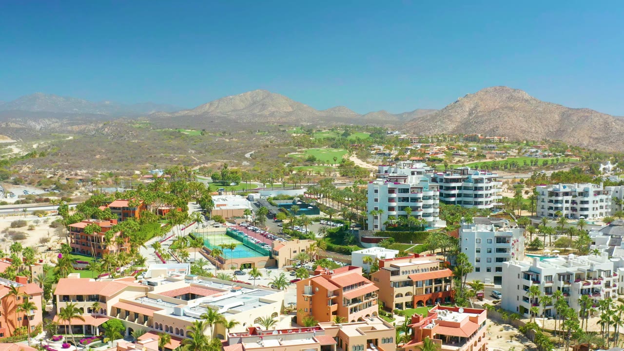 Opening aerial frame over Dreams Los Cabos in Baja California Sur, showing white and terracotta buildings, pools, palm trees, golf course greens, dry hills, mountains, and no visible people.
