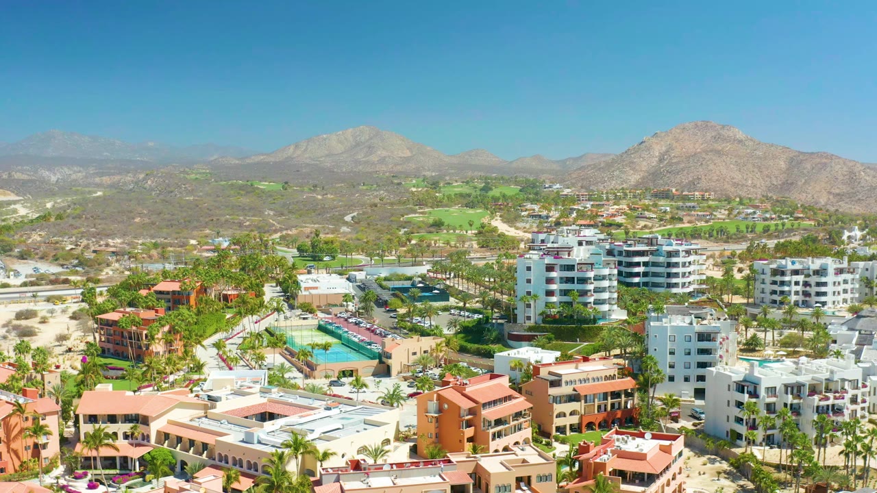 Aerial frame at Dreams Los Cabos with a slightly advanced view over white apartment-style buildings, turquoise pools, palm-lined roads, green fairways, mountains, and no visible people.