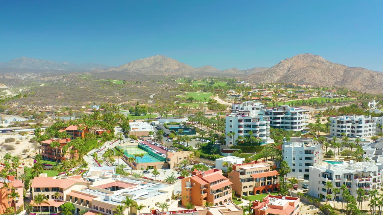 Aerial scene with pools and resort buildings in front of golf course greens and mountains.