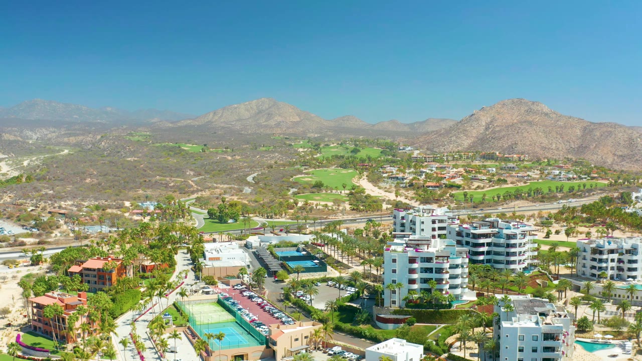 The second frame keeps a wide aerial angle over Dreams Los Cabos, with apartment blocks and pools in front, bright golf fairways beyond, rocky hills, distant mountains, and no visible people.