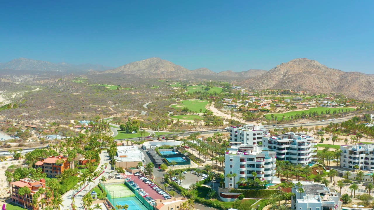 A wide aerial frame over Dreams Los Cabos shows foreground residential blocks and parking areas, curved roads, green golf course sections, scrub-covered hills, mountain ridges, and no visible people.