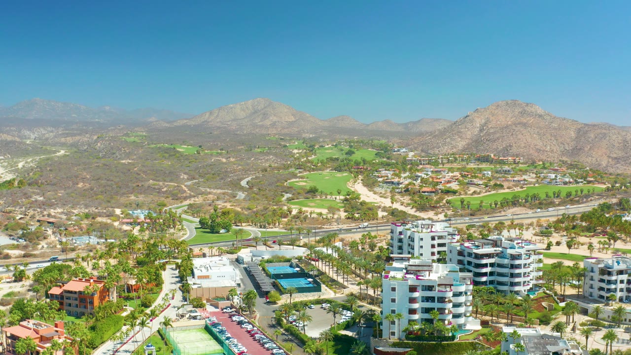 Aerial view with white buildings, pool deck, palms, road, golf course, and mountains