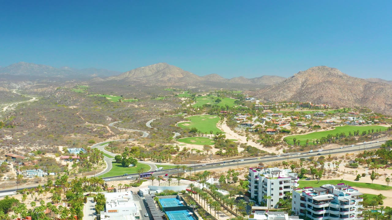 Aerial view over Dreams Los Cabos in Baja California Sur with white foreground buildings, a curving highway, green golf fairways, dry hills, distant mountains, and no visible people.