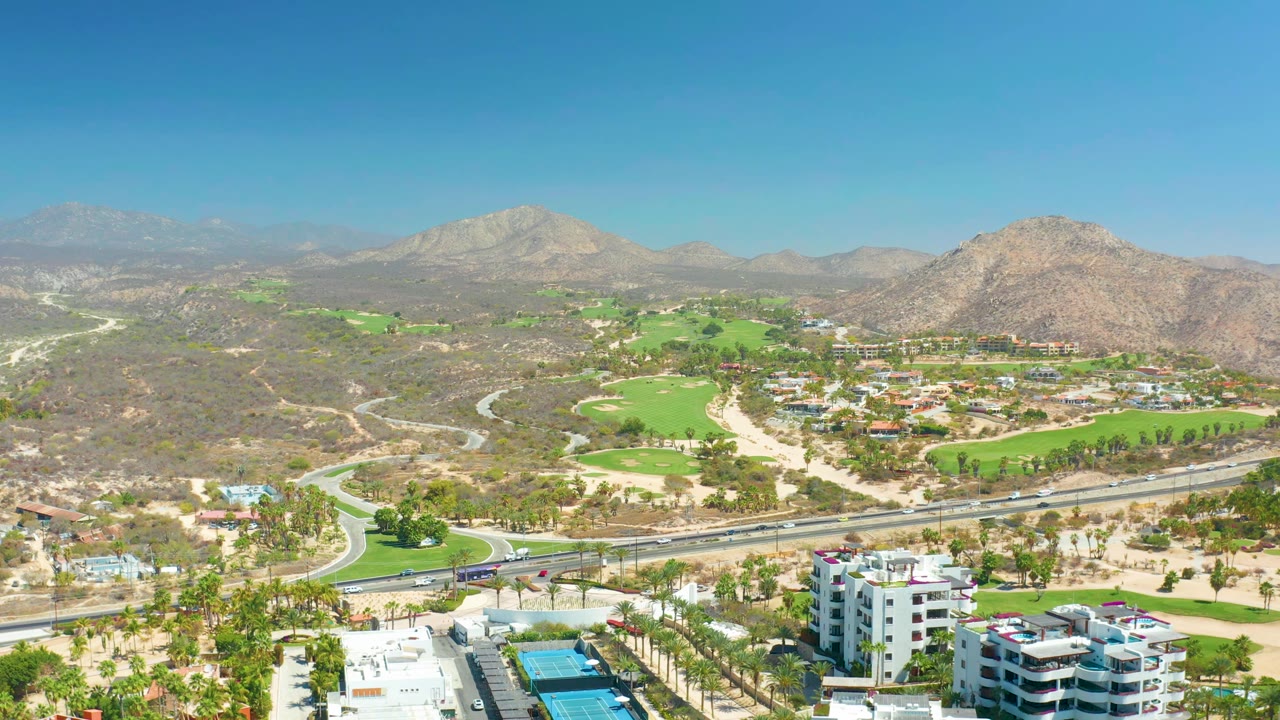 Wide aerial over green fairways, road traffic, palms, dry hills, and distant mountains.