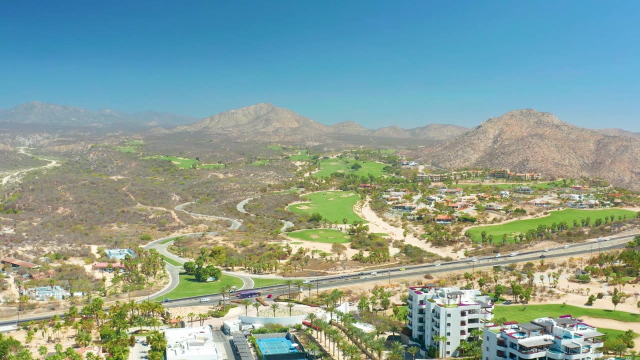 Aerial view in Baja California Sur with the golf course centered, a paved highway and cars along the lower frame, low resort-area buildings, dry slopes, mountains, and no visible people.