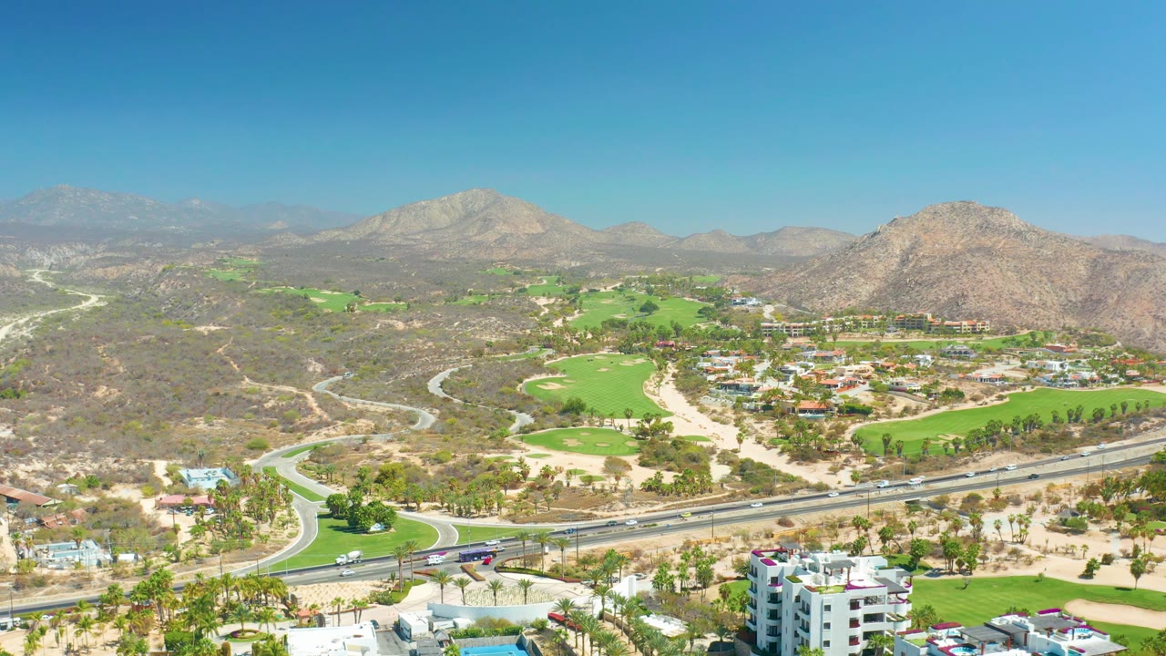 Aerial landscape with golf greens, winding road, desert hills, and mountain horizon.