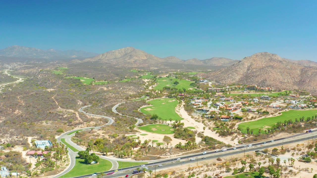 Opening aerial frame at Dreams Los Cabos shows bright golf fairways beside a highway, dry scrub terrain, rocky mountains, and no visible people.