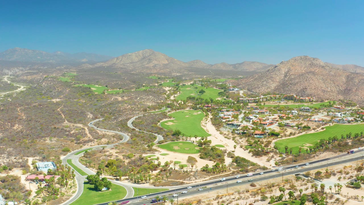 At 0:02, the wide Dreams Los Cabos frame shows the golf course and nearby buildings set between arid hills, foreground highway lanes, and no visible people.