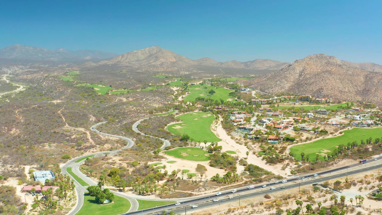 At 0:03, Dreams Los Cabos appears from above with fairways, sand-colored course areas, a busy highway foreground, rocky hills, and no visible people.