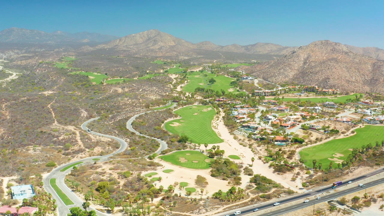 Opening aerial frame at Dreams Los Cabos shows green fairways, sand bunkers, curving roads, scattered buildings, dry hills, distant mountains, and no visible people.