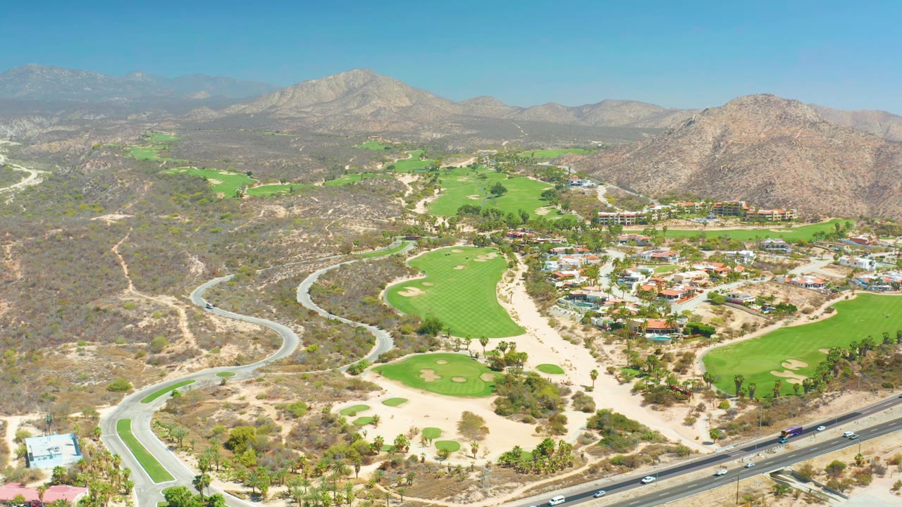At 0:01 the elevated view holds on Dreams Los Cabos golf greens, a looping road at left, small traffic on the lower highway, buildings near the fairways, and no visible people.