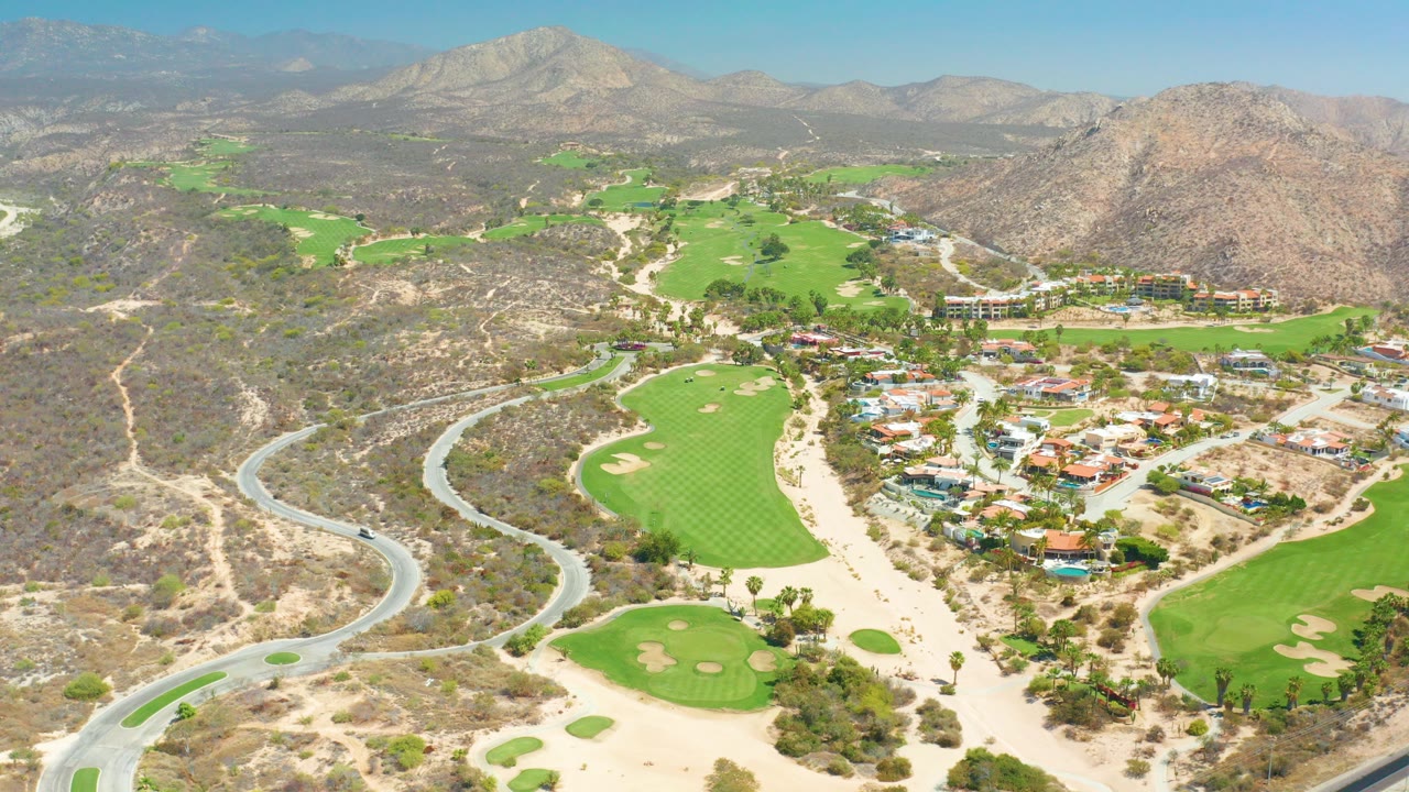 Opening aerial frame over Dreams Los Cabos showing bright golf fairways, a curving road at left, low buildings, dry hills, rocky mountains, and no visible people.