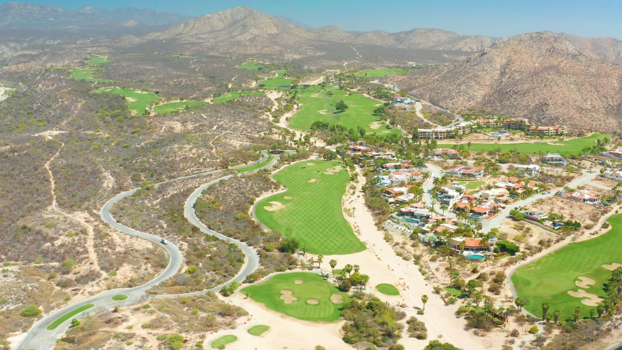 Drone-height frame over Dreams Los Cabos where the car continues along the curved road at left, fairways fill the lower center, buildings sit at right, and no visible people appear.