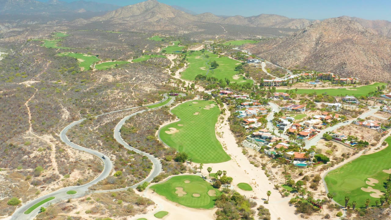 Wide aerial of green fairways, desert slopes, buildings, and winding road.