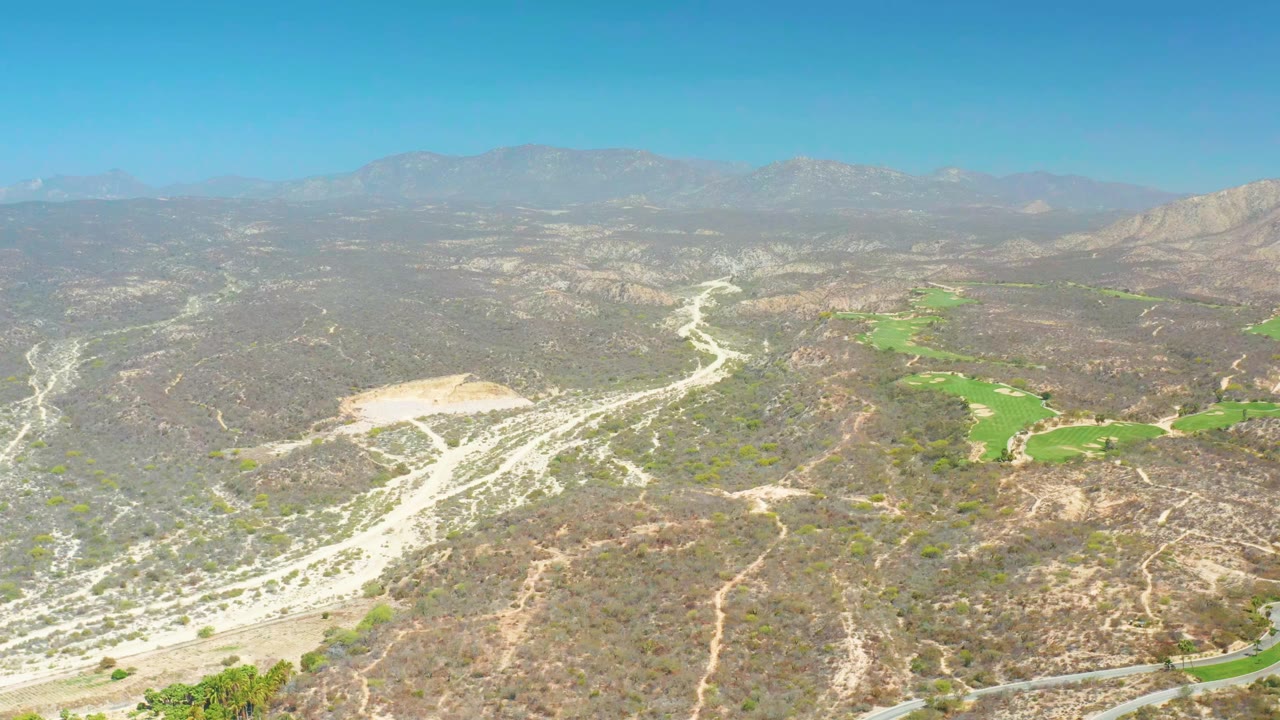 Wide drone-style view at Dreams Los Cabos with pale dry washes crossing scrub-covered hills, green fairways on the right side, hazy mountain ridges, and no visible people.