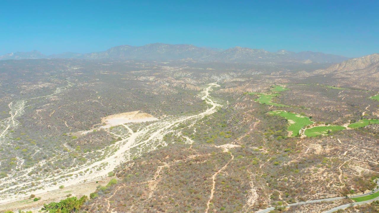 Aerial view over Dreams Los Cabos terrain showing white riverbed channels, brown hills, golf course greens to the right, a road near the lower edge, mountains, and no visible people.