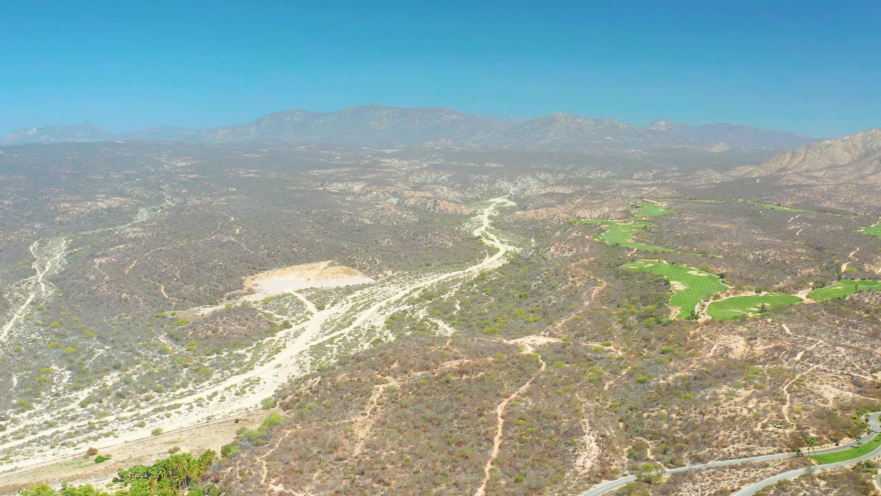 At 0:01, the Dreams Los Cabos aerial view holds on tan hills, branching pale tracks, a curving road near the lower edge, green patches on the right, distant mountains, and no visible people.