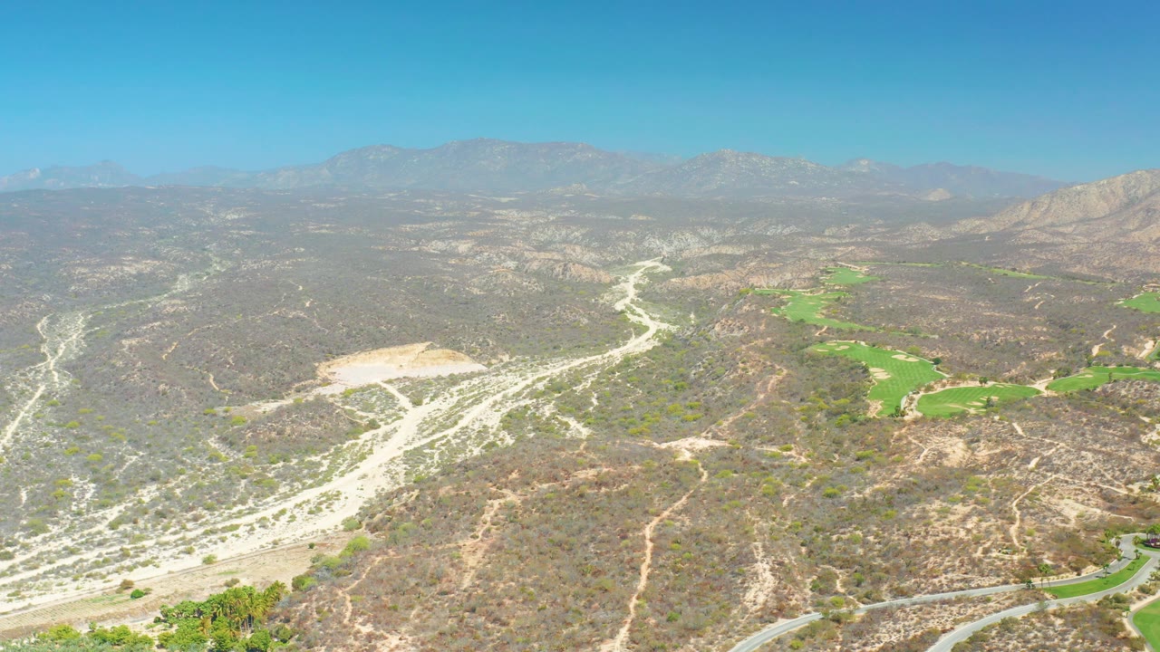 At 0:03, the Dreams Los Cabos frame presents a broad aerial composition with pale wash lines cutting through dry hills, green patches on the right side, distant mountains, clear blue sky, and no visible people.