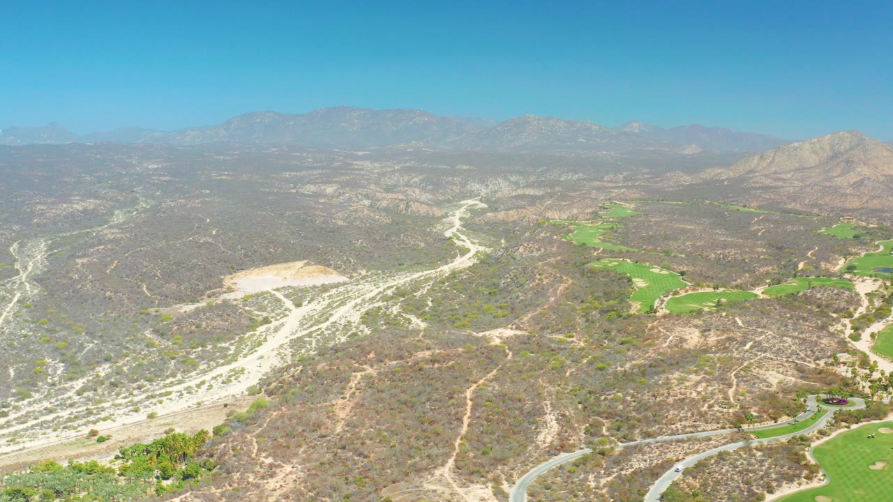 At Dreams Los Cabos in Baja California Sur, an aerial view looks over dry hills, pale winding roads or washes, green golf-course patches, distant mountains, clear blue sky, and no visible people.
