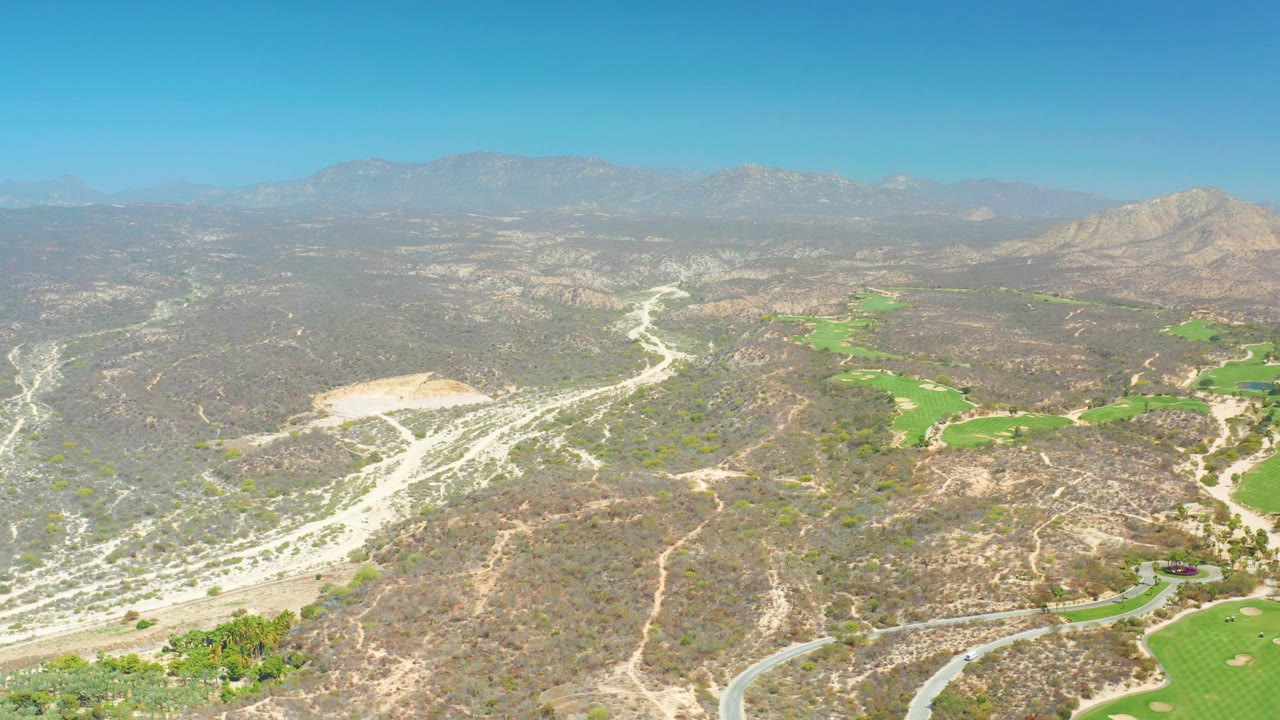 At 0:02, the aerial view over Dreams Los Cabos shows dry ridges in the foreground, a curving paved-looking road at the lower edge, green course patches to the right, distant mountains, and no visible people.