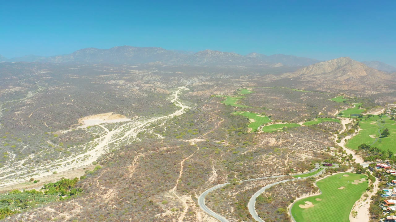 At 0:02, the wide aerial composition shows the golf course lower right, several curved road lines through the dry hills, layered mountains in the background, clear sky, and no visible people.