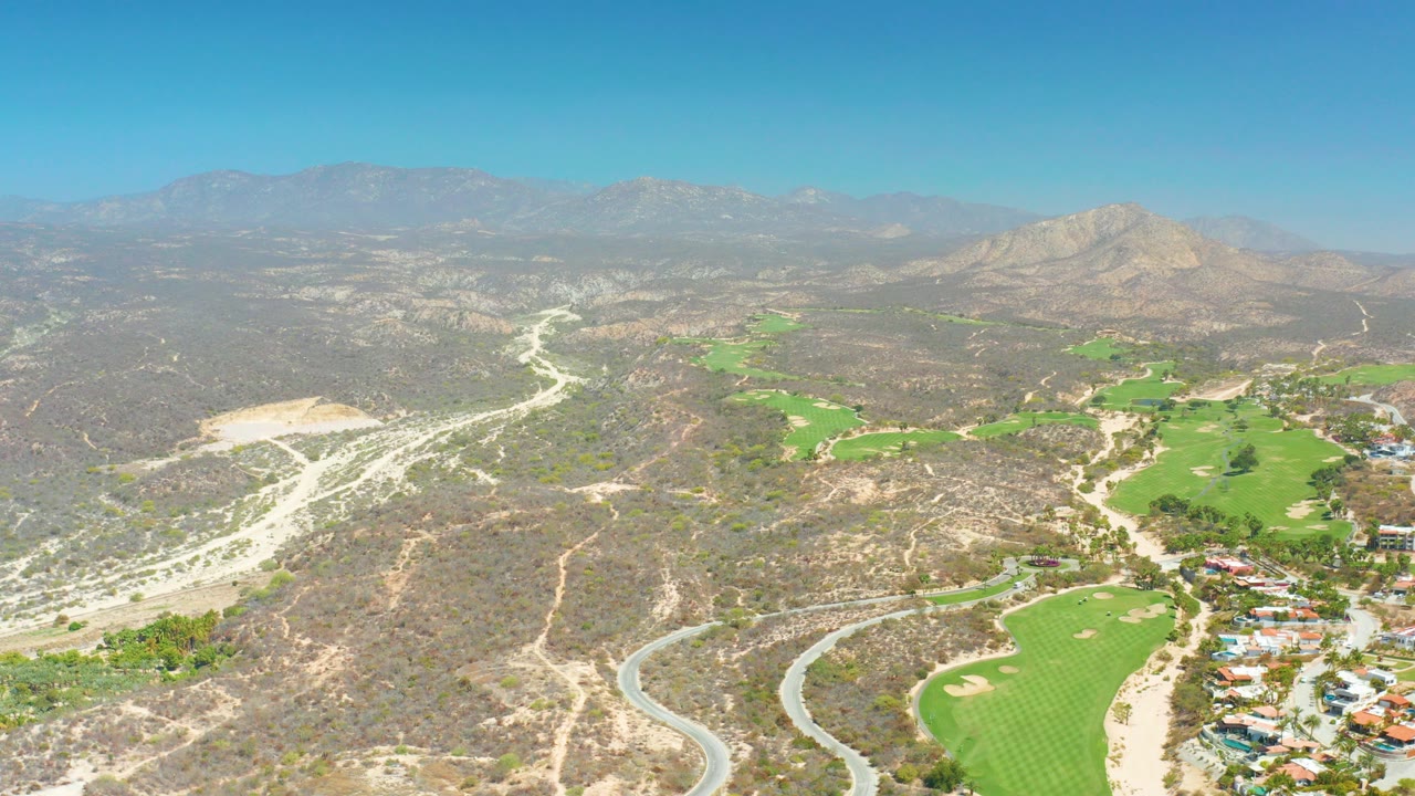 Opening aerial frame over Dreams Los Cabos, Baja California Sur, showing a winding pale road through dry hills, green golf course areas, scattered buildings, distant mountains, clear blue sky, and no visible people.