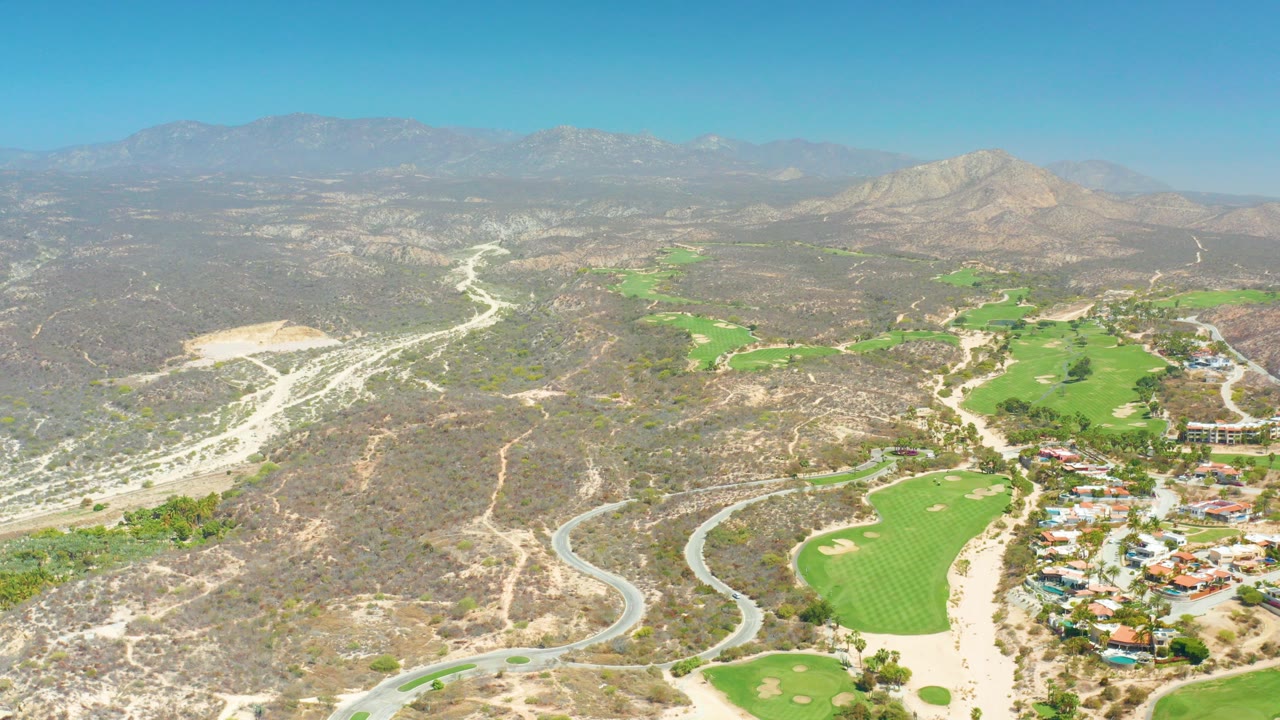 Aerial frame over Dreams Los Cabos showing a winding road at lower left, bright green fairways in the valley, dry hills, distant mountains, clear blue sky, and no visible people.