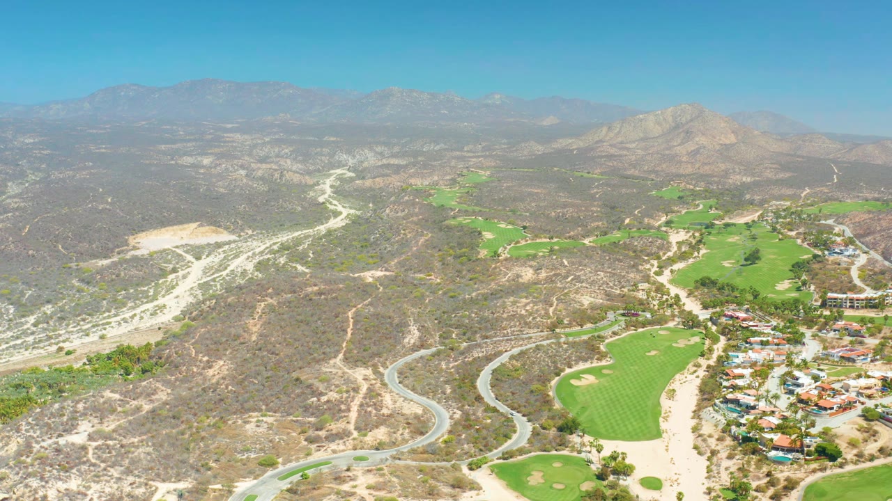 Second aerial frame at Dreams Los Cabos with the curving road lower in frame, clustered buildings near the fairways, brown scrub hills, mountain ridges, blue sky, and no visible people.