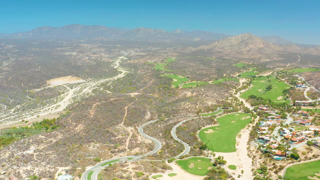 Aerial view over Dreams Los Cabos with pale dirt roads crossing dry terrain, green course sections through the middle, buildings to the right, hazy mountains, and no visible people.