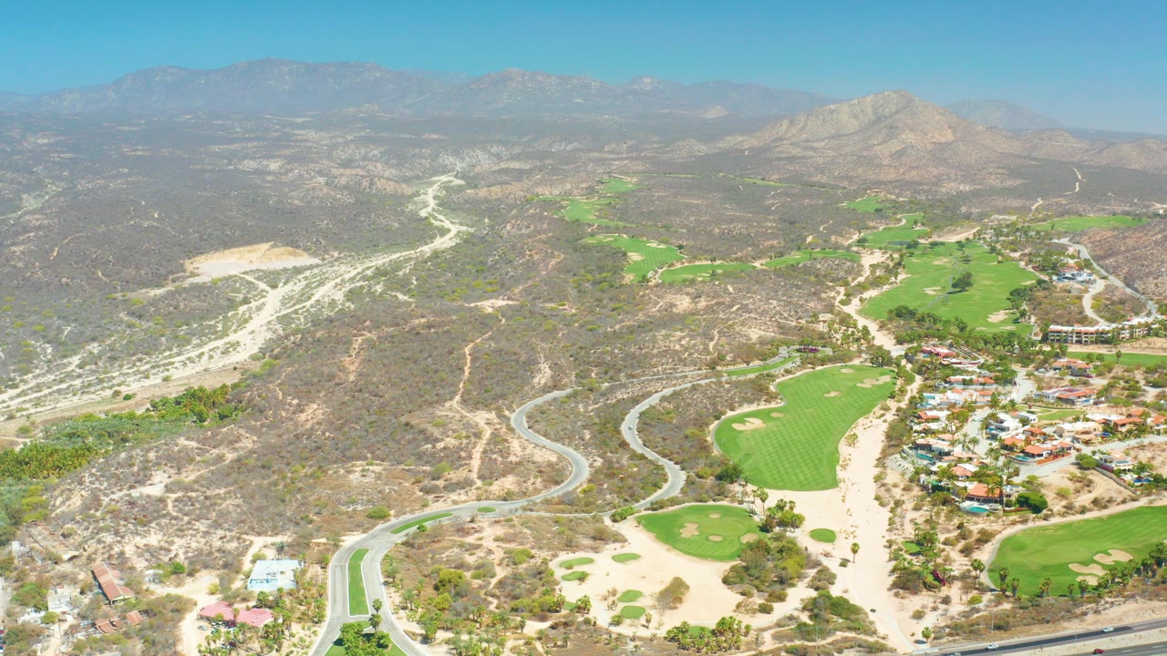 Opening aerial frame over Dreams Los Cabos with a winding road, bright golf greens, dry hills, distant mountains, blue sky, and no visible people.
