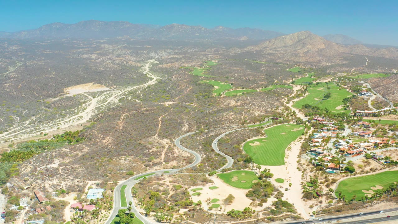Aerial frame over Dreams Los Cabos with golf greens and sand traps in the lower area, a winding road with small vehicles, dry hills, mountain horizon, and no visible people.