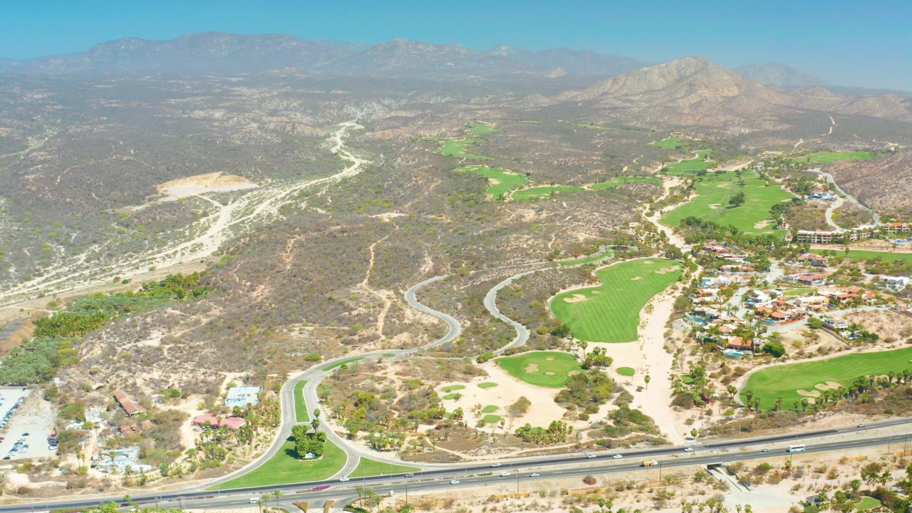 High aerial view over Dreams Los Cabos, Baja California Sur, with a divided highway at the bottom, green golf fairways in the middle distance, dry hills, distant mountains, and no visible people.