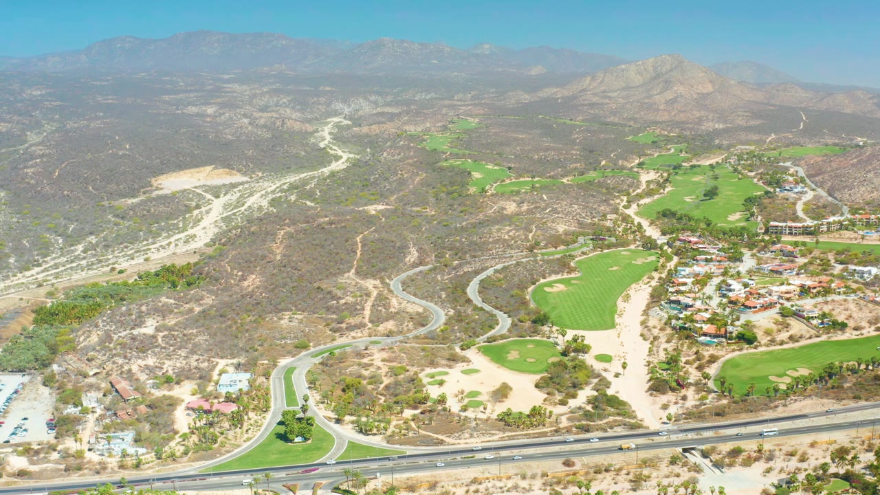 Drone-height frame in Dreams Los Cabos showing the highway and roundabout near the lower edge, bright golf greens through the center, scrub-covered hills, pale roads, mountains, and no visible people.