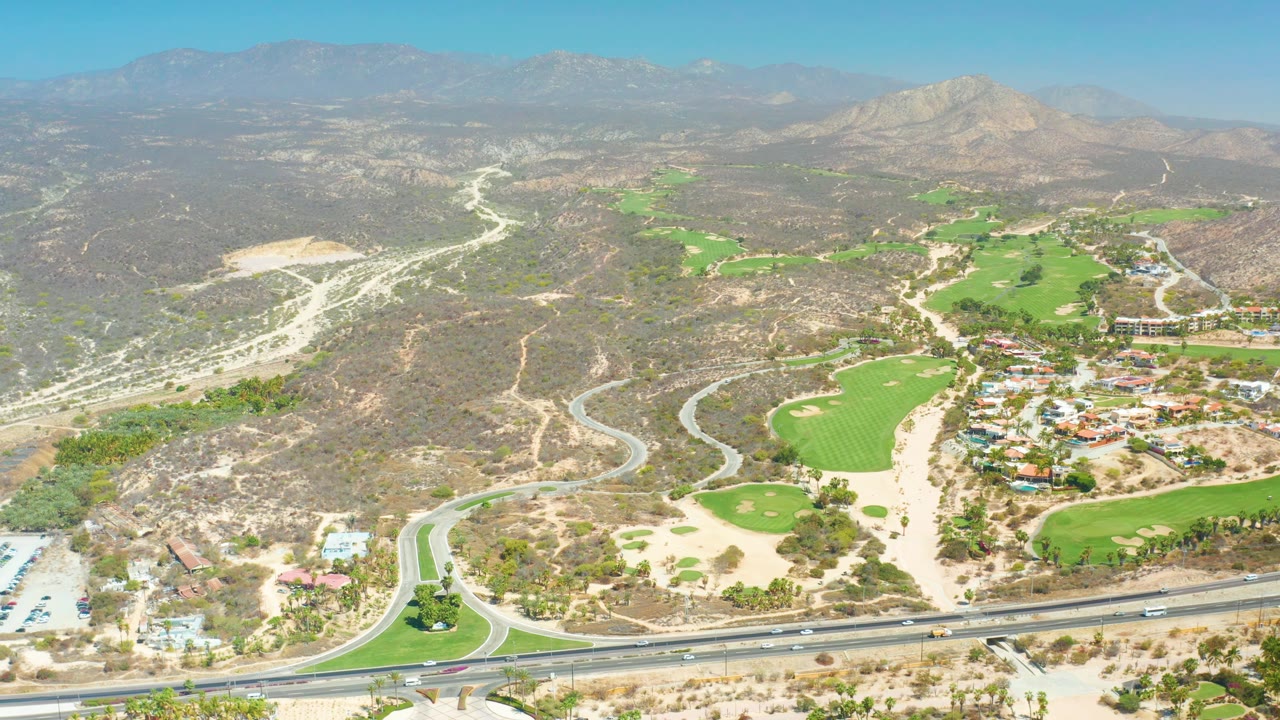 Aerial frame over Dreams Los Cabos with small vehicles on the lower highway, resort-area buildings beside green fairways, pale dry hills behind them, mountain ridges on the horizon, and no visible people.