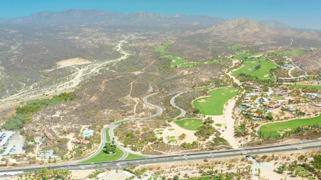 Wide aerial view in Dreams Los Cabos, Baja California Sur, with the divided highway across the foreground, golf course greens and sand-colored paths in the midground, dry hills, mountains, and no visible people.
