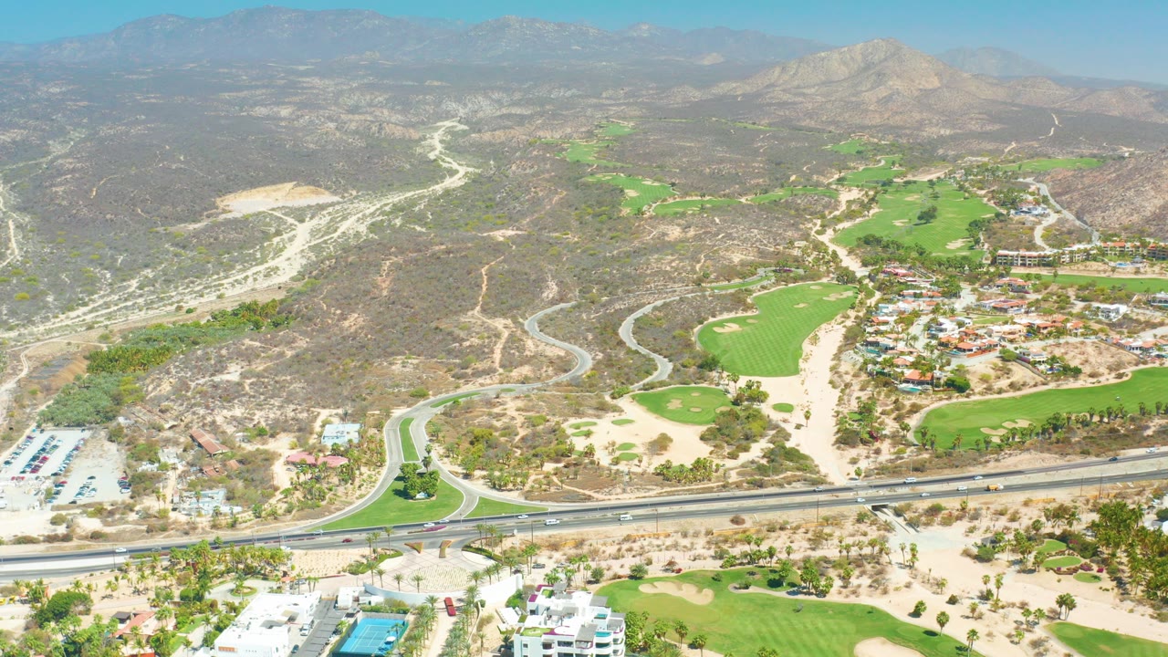 Aerial view of highway, golf course, dry hills, resort buildings, and distant mountains.