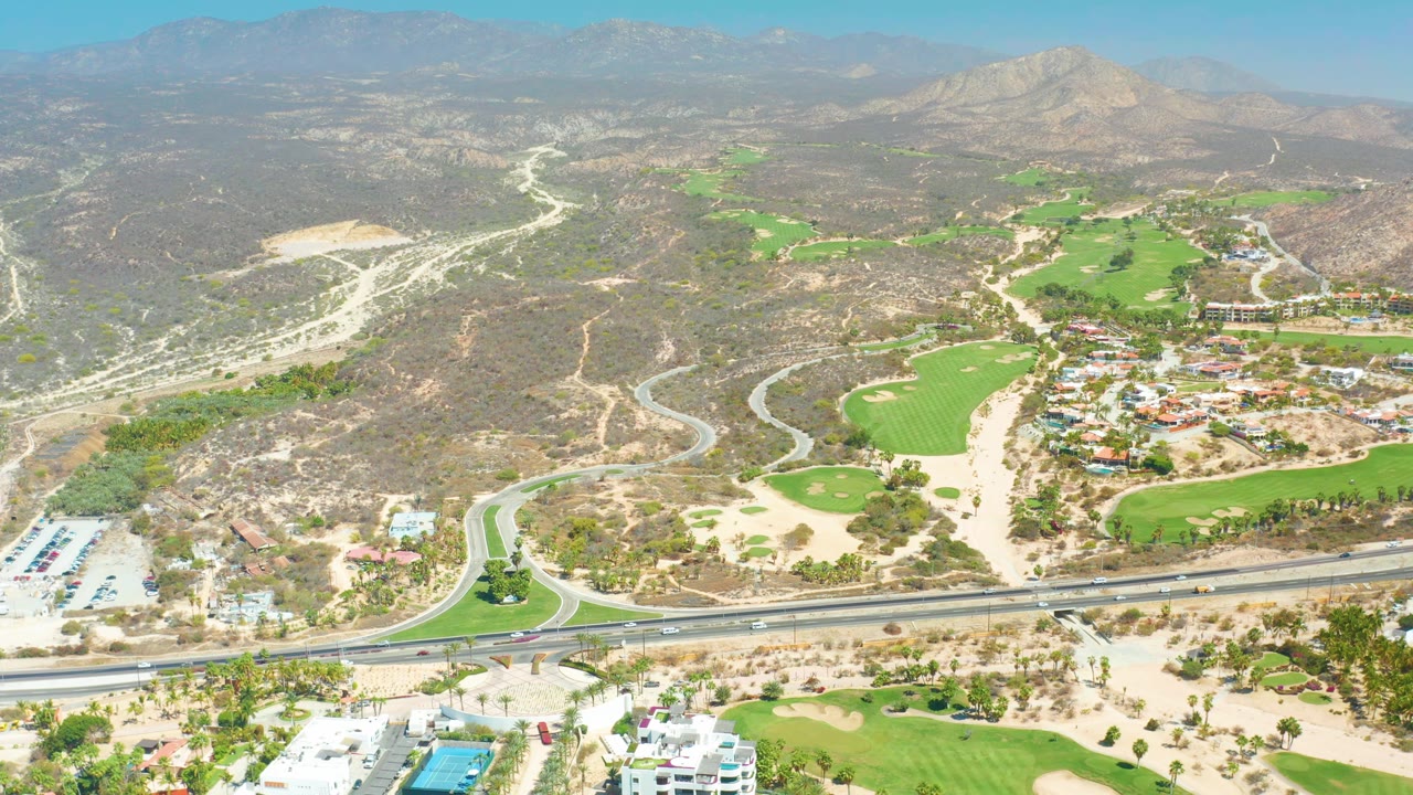 Aerial frame at Dreams Los Cabos with the roundabout and highway lower left, golf greens running up the valley, resort buildings to the lower right, mountains beyond, and no visible people.