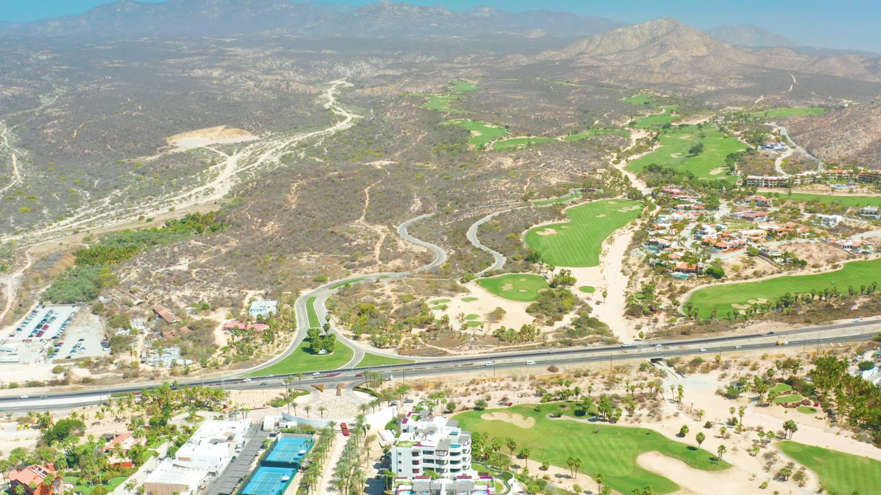 Dreams Los Cabos aerial frame with the roundabout near the lower left, highway across the foreground, golf course and tan paths in the center, dry hills in the background, and no visible people.