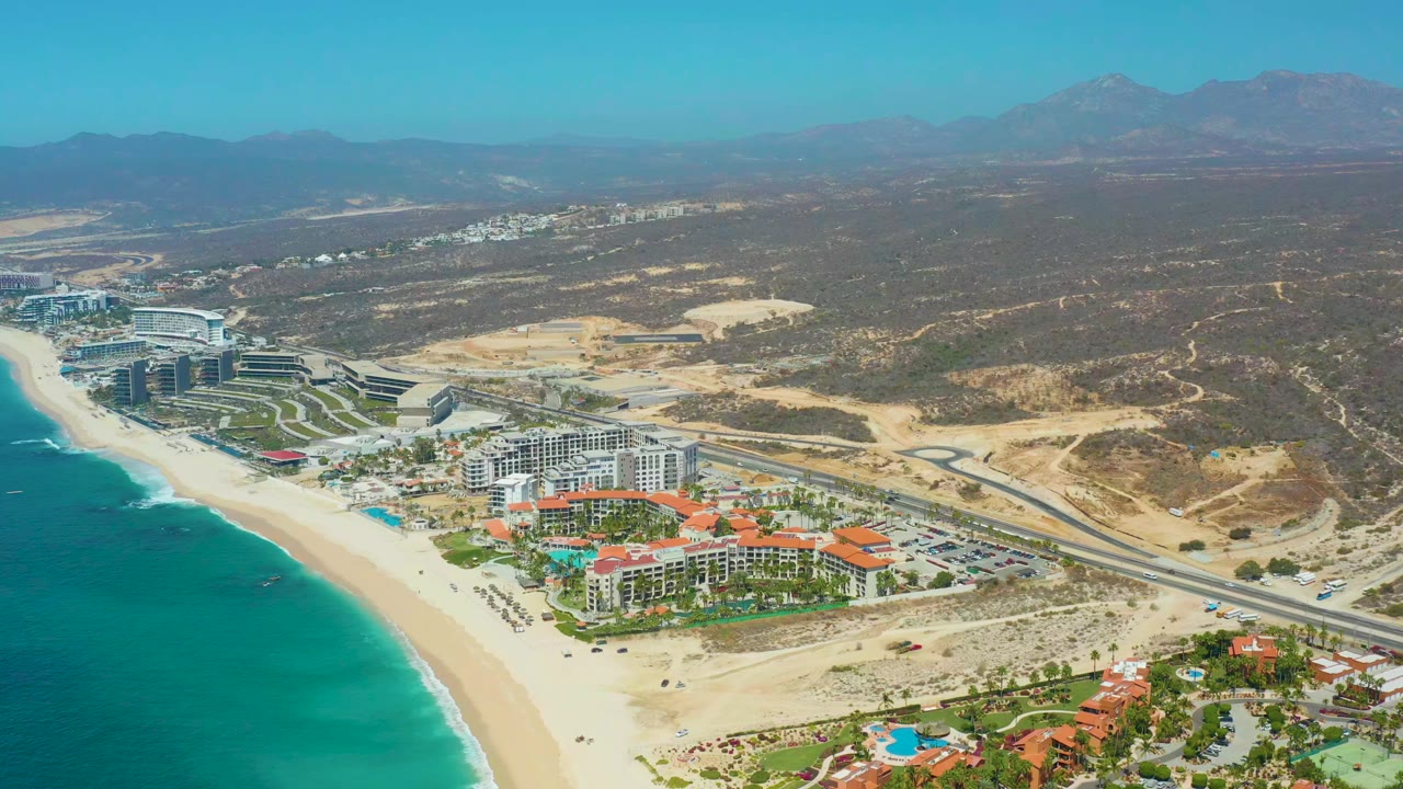 Opening aerial frame over Dreams Los Cabos with turquoise water at left, a wide sandy beach, resort buildings near the shore, a road behind them, desert scrubland, distant mountains, and tiny beachgoers visible.