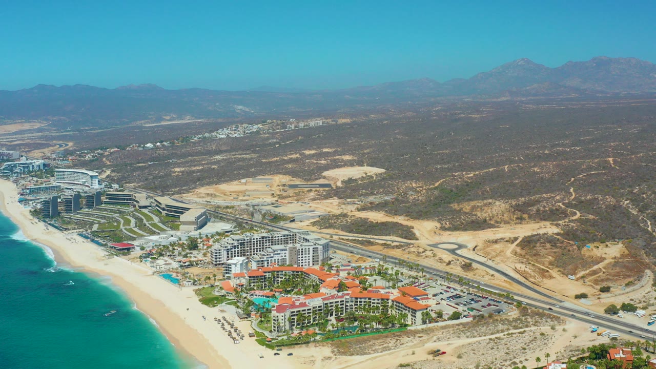 Wide aerial frame over Dreams Los Cabos, Baja California Sur, with turquoise ocean at left, sandy beach, resort buildings near the shore, a coastal road, dry hills, distant mountains, and no visible people.