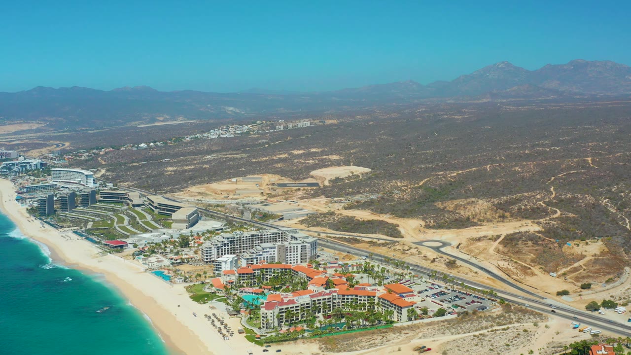 High drone frame over Dreams Los Cabos with pale sand and blue water on the left, white and terracotta-roof buildings near the shore, a multi-lane coastal road, open dry lots, hills, mountains, and no visible people.
