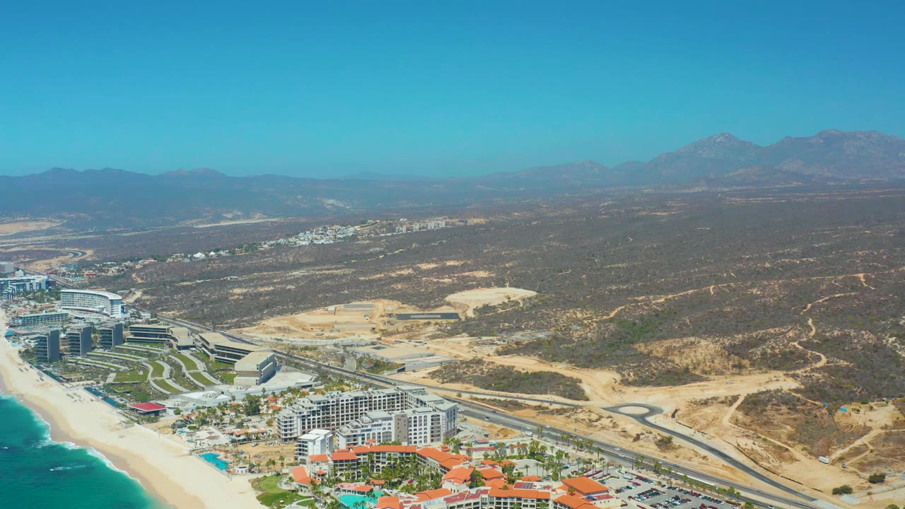 Wide aerial view at Dreams Los Cabos showing the coastline, resort complex, roadside development, dry scrubland, and mountain ridge under a clear sky, with no visible people.