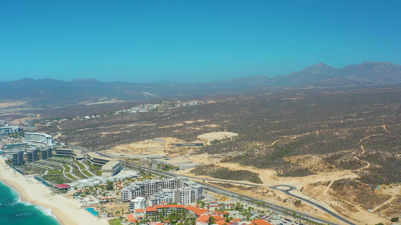 High aerial frame from the Dreams Los Cabos coastline showing oceanfront buildings and beach in the lower left, a road cutting through dry hills, and mountains in the background, with no visible people.