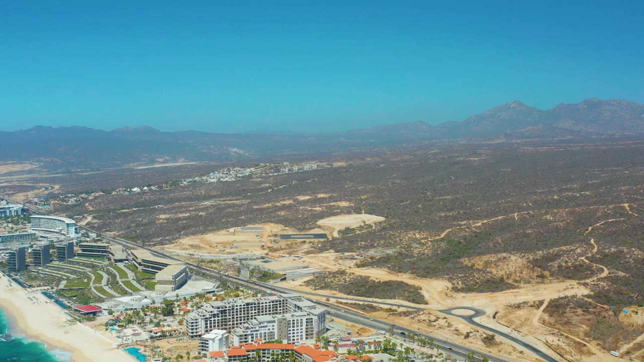 Aerial frame over Dreams Los Cabos showing beachfront development low in frame, pale sandy road areas through the middle, broad arid hills, distant mountains, and no visible people.