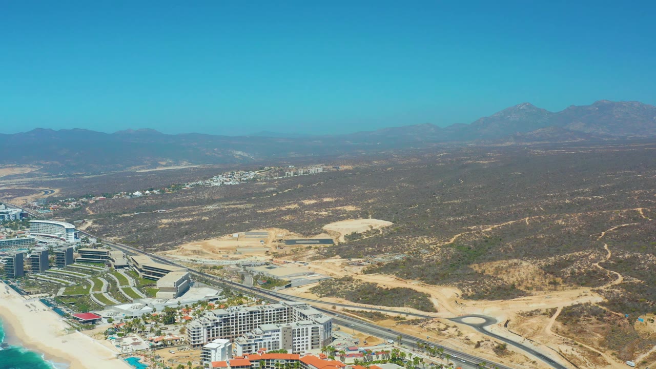 Dreams Los Cabos aerial view with ocean color at the lower left edge, resort structures and a road junction below, open dry terrain behind them, mountain ridges, and no visible people.