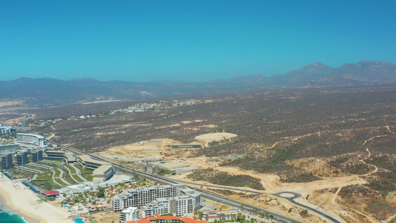 Mid-clip aerial frame at Dreams Los Cabos with resort buildings partly at the bottom, a curved road and pale earthworks in the center, low arid hills, distant mountains, and no visible people.