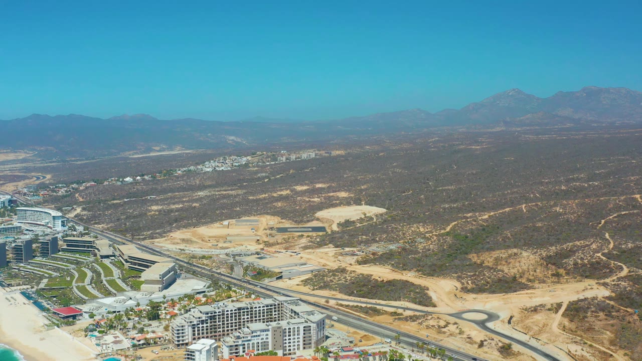 Aerial frame at Dreams Los Cabos showing the resort structures slightly left of center, a paved road through graded sandy land, mountain ridges, and no visible people.